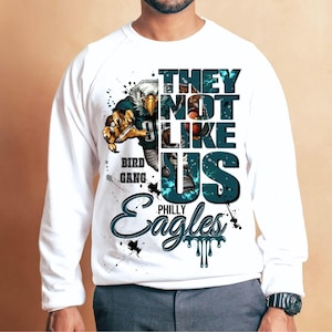 Może przedstawiać: Biała bluza z grafiką orła w stroju futbolowym. Tekst brzmi "THEY NOT LIKE US" i "PHILLY Eagles". Projekt zawiera słowa "BIRD GANG" i efekty rozprysk&oacute;w.