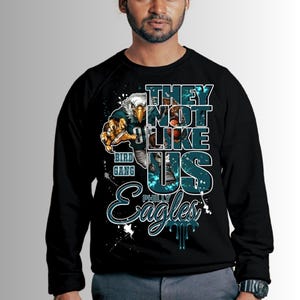 Może przedstawiać: Czarna bluza z okrągłym dekoltem z graficznym wzorem przedstawiającym orła w stroju futbolowym. Tekst brzmi "THEY NOT LIKE US" i "PHILLY Eagles" w kolorze turkusowym i białym. Projekt zawiera plamy farby.