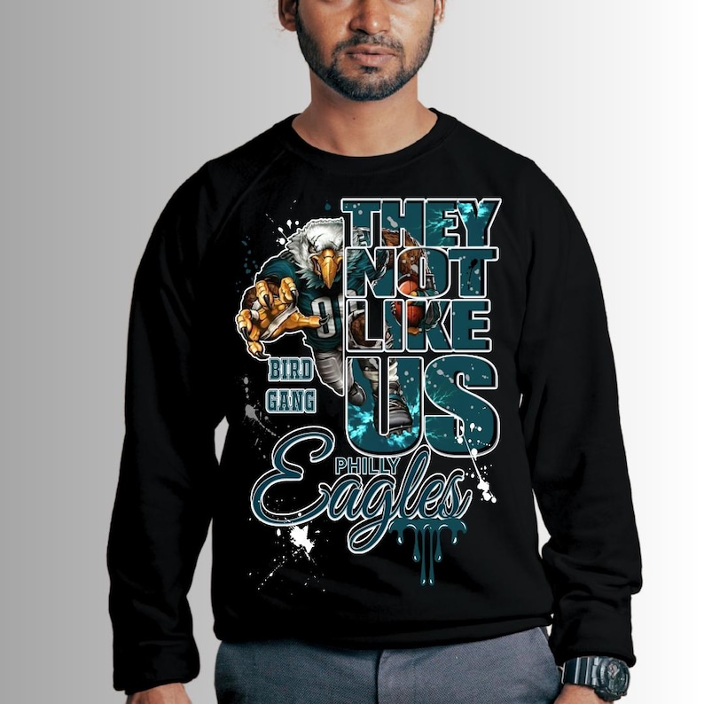 Może przedstawiać: Czarna bluza z okrągłym dekoltem z graficznym wzorem przedstawiającym orła w stroju futbolowym. Tekst brzmi "THEY NOT LIKE US" i "PHILLY Eagles" w kolorze turkusowym i białym. Projekt zawiera plamy farby.
