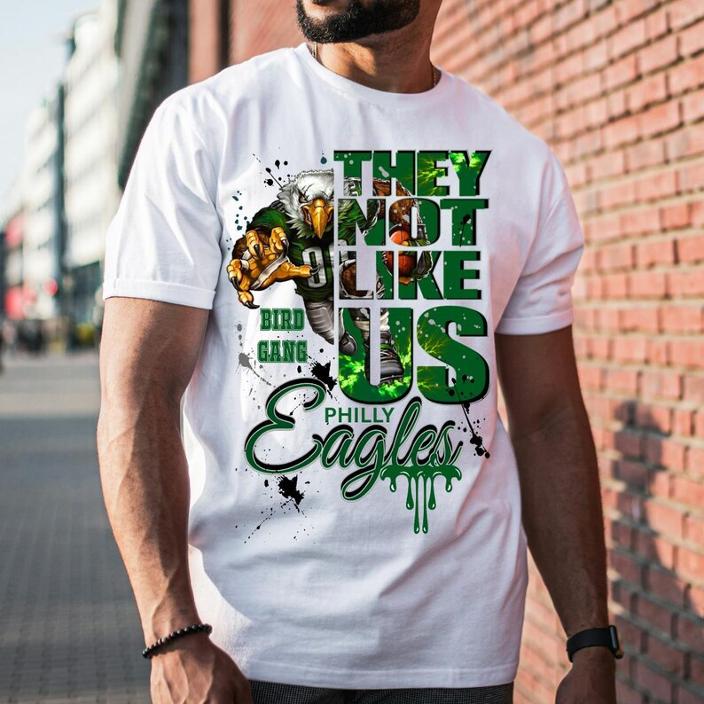 Może przedstawiać: Biała koszulka z zielono-czarnym nadrukiem graficznym. Projekt przedstawia orła, tekst "THEY NOT LIKE US", "BIRD GANG" i "PHILLY Eagles". Koszulka ma okrągły dekolt i kr&oacute;tkie rękawy.
