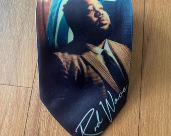 May include: A necktie featuring a photographic print of a person wearing a tan and brown patterned blazer with a white shirt and black tie. The lower portion of the tie is black and displays the white cursive text Rod Wave printed on the fabric.