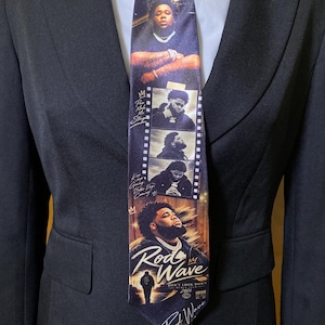 May include: A black necktie featuring a graphic print of the artist Rod Wave. The design includes multiple photographic portraits of the artist, film strip elements, and text that reads Rod Wave, Don't Look Down, and World Tour 2024.