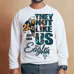 Może przedstawiać: Biała bluza z nadrukiem przedstawiającym drapieżnego orła oraz napisy "THEY NOT LIKE US" i "PHILLY EAGLES". Projekt zawiera kolory morski i czarny, widoczne są r&oacute;wnież słowa "BIRD GANG".