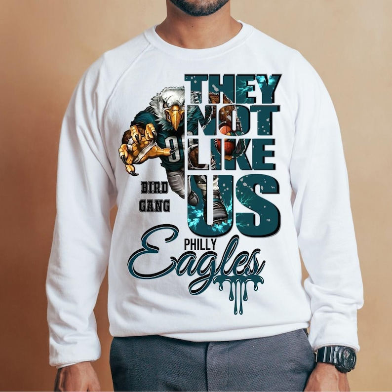 Może przedstawiać: Biała bluza z nadrukiem przedstawiającym drapieżnego orła oraz napisy "THEY NOT LIKE US" i "PHILLY EAGLES". Projekt zawiera kolory morski i czarny, widoczne są r&oacute;wnież słowa "BIRD GANG".