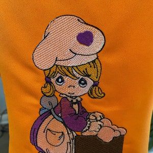 May include: An orange fabric background featuring an embroidered design of a cartoon character with blonde hair, wearing a purple shirt and a light-colored hat with a purple heart. The character is sitting next to a brown rectangular object.