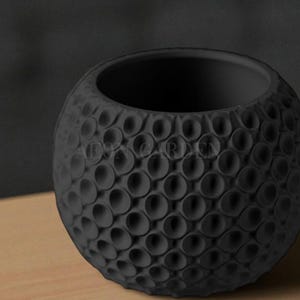 May include: A black, spherical ceramic planter with a textured, dimpled surface. The planter has a wide opening and sits on a light-colored wooden surface. The background is a dark gray.