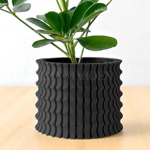 May include: A black, textured plant pot with a wavy, vertical design. The pot holds a green plant with large, rounded leaves. The pot is sitting on a light-colored wooden surface.
