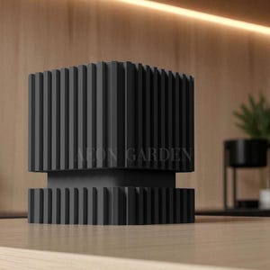 May include: A black, modern, geometric sculpture with vertical ridged detailing. The sculpture has a rectangular base and a larger, ridged top section. The text "AEON GARDEN" is visible on the front. The sculpture is displayed on a light wood surface.