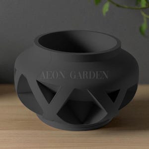 May include: A dark gray, decorative ceramic planter with a geometric lattice design. The planter has a round shape with a wide opening and the text "AEON GARDEN" is printed on the side. The planter is sitting on a wooden surface.