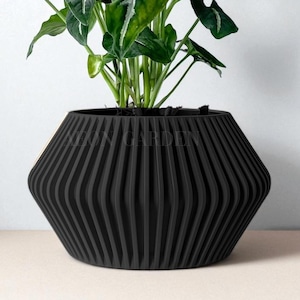 May include: A black, geometric-shaped planter with vertical ridged detailing. The planter holds a green plant with large, veined leaves. The planter is sitting on a light-colored surface against a white background. The text "KEVIN GARDEN" is visible on the planter.