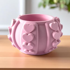 May include: A pink planter with a rounded shape and heart-shaped embellishments. The planter has a wide opening and is made of a solid material. The hearts are evenly spaced around the planter's exterior.