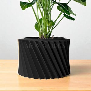 May include: A black, geometric planter with a spiral design, holding a green leafy plant. The planter has the text "AEON GARDEN" on the side. The planter sits on a light brown wooden surface.