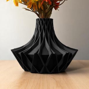 May include: A black geometric vase with a diamond pattern, holding a bouquet of colorful artificial flowers. The vase has a wide base and a narrow neck. The flowers include yellow, orange, and red blooms.