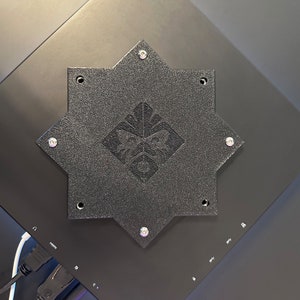 May include: A black, octagonal-shaped metal plate with a logo in the center. The plate is attached to a black surface with screws.
