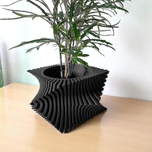 May include: A black, geometric planter with a unique, wave-like design. The planter holds a green leafy plant. The planter is made of a solid material and sits on a light-colored wooden surface. The design is modern and eye-catching.
