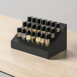 May include: A black, angled display stand holding numerous small glass bottles with black caps. The bottles contain various shades of liquid, ranging from clear to amber. The stand is on a light-colored wooden surface.