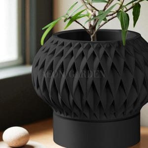 May include: A black, geometric-patterned planter pot with a small plant inside. The pot has a textured, diamond-like design and a solid base. The text "AEON GARDEN" is visible on the pot. A small, round, beige stone sits on a wooden surface.