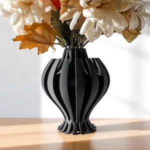 May include: A black, modern vase with a unique, slatted design. The vase is filled with a bouquet of cream and orange-toned artificial flowers and foliage. The vase sits on a light brown wooden surface.