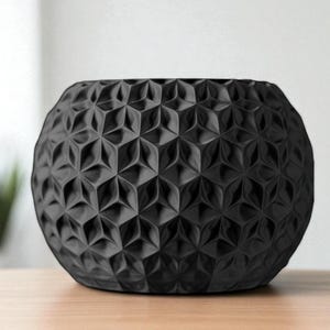 May include: A black, spherical decorative bowl with a geometric, textured pattern. The bowl is made of a matte material and sits on a light-colored wooden surface. The bowl's design features repeating three-dimensional shapes.