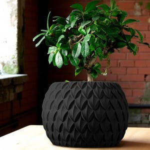 May include: A small bonsai tree with vibrant green leaves sits in a black, textured, round planter. The planter has a repeating, geometric pattern. The background features a brick wall and a window.