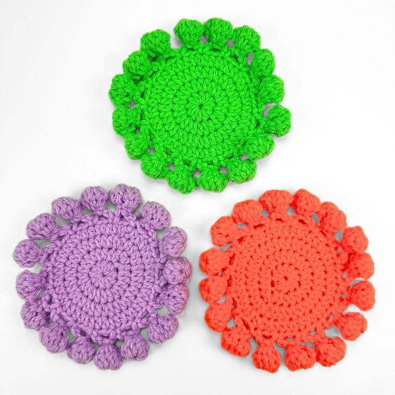 May include: Three circular crochet coasters in bright neon green, soft lavender, and vibrant orange. Each coaster features a dense, textured center with a decorative border of small, rounded bobble stitches around the entire outer edge.