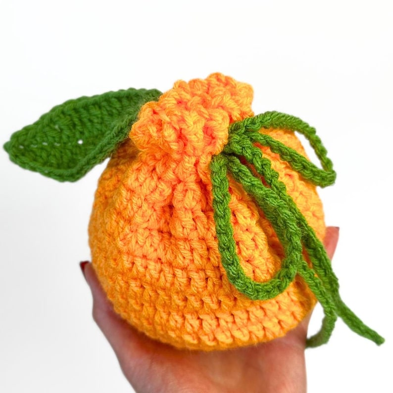 K&ouml;nnte beinhalten: Ein runder, orangefarbener H&auml;kelbeutel mit Kordelzug in Form einer Zitrusfrucht. Der Beutel hat ein strukturiertes Maschenbild, ein einzelnes gro&szlig;es gr&uuml;nes H&auml;kelblatt an der Seite und einen gr&uuml;nen H&auml;kelkordelzug als Verschluss.