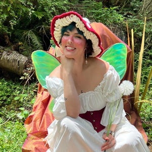 May include: A crochet mushroom hat with a red cap and white ruffled brim, paired with a white off-the-shoulder dress, a dark red corset-style bodice, and iridescent green fairy wings. The ensemble features a whimsical aesthetic with natural elements.