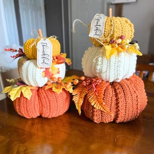 Puede incluir: Dos conjuntos apilados de tres calabazas de ganchillo en naranja, blanco y amarillo. Las calabazas est&aacute;n decoradas con hojas de oto&ntilde;o artificiales, bayas y tallos de madera. Una etiqueta a la izquierda dice Thankful y a la derecha Grateful.