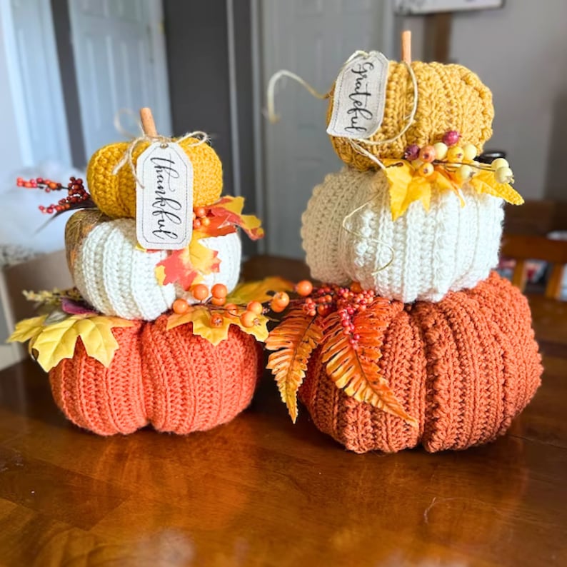 Puede incluir: Dos conjuntos apilados de tres calabazas de ganchillo en naranja, blanco y amarillo. Las calabazas est&aacute;n decoradas con hojas de oto&ntilde;o artificiales, bayas y tallos de madera. Una etiqueta a la izquierda dice Thankful y a la derecha Grateful.