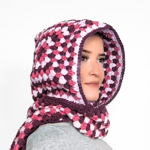May include: A crochet hood with a granny square pattern in shades of dark purple, magenta, and light pink. The hood features a solid dark purple border along the front opening and extends into a scarf-like section that wraps around the neck area.