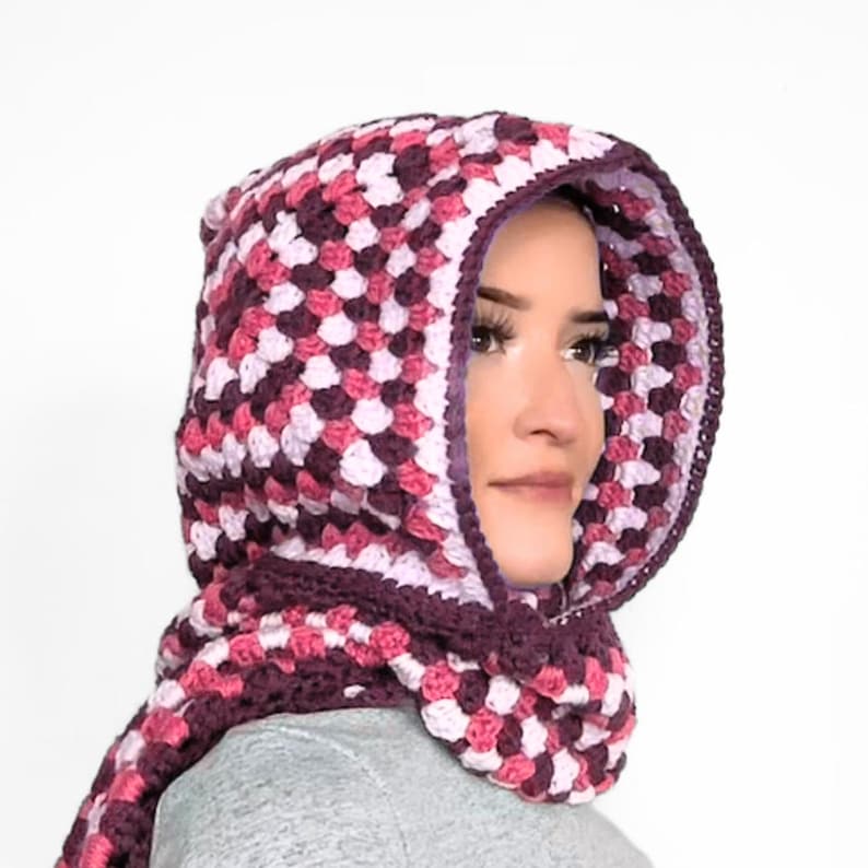 May include: A crochet hood with a granny square pattern in shades of dark purple, magenta, and light pink. The hood features a solid dark purple border along the front opening and extends into a scarf-like section that wraps around the neck area.