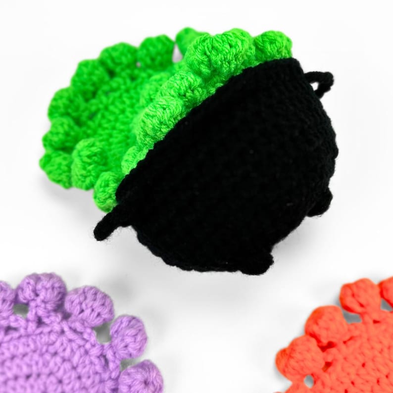 May include: A black crocheted cauldron with a bright green textured top, accompanied by two additional crocheted circular coasters in solid purple and solid orange, each featuring a scalloped edge design made of small yarn loops.