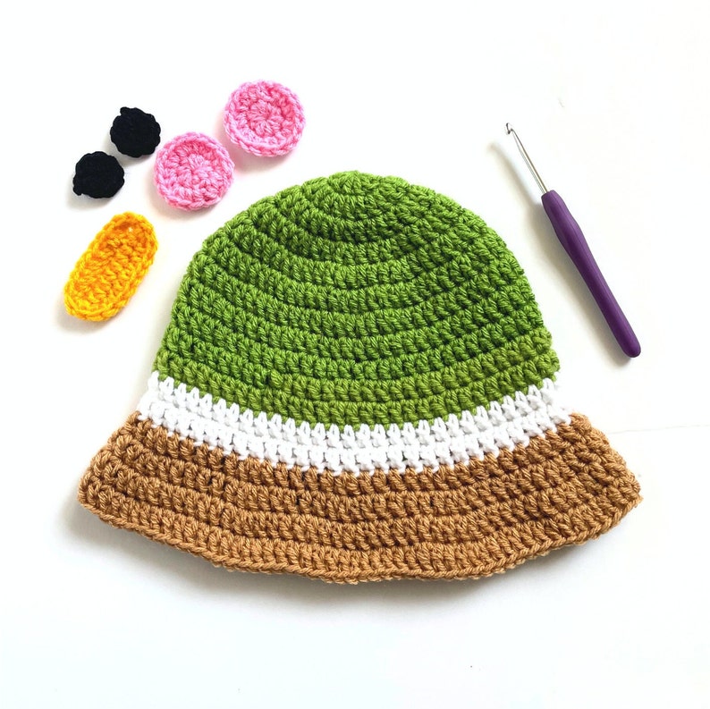Pu&ograve; includere: Un cappello a secchiello di uncinetto verde, bianco e marrone con un uncinetto e decorazioni in feltro colorato.