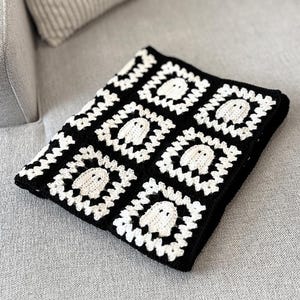 May include: A square crochet blanket featuring a repeating pattern of white ghost motifs centered within black and white granny squares. The textile is constructed from interconnected yarn squares, each displaying a small, stylized ghost figure.
