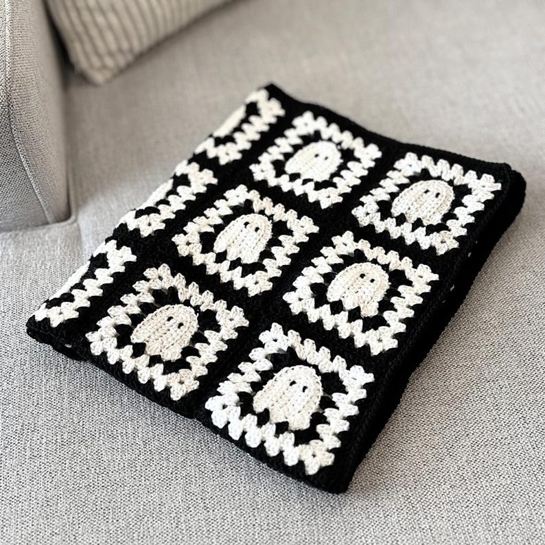 May include: A square crochet blanket featuring a repeating pattern of white ghost motifs centered within black and white granny squares. The textile is constructed from interconnected yarn squares, each displaying a small, stylized ghost figure.