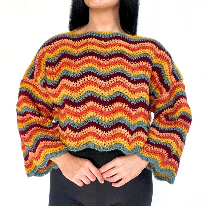 May include: A long-sleeve crochet sweater featuring a horizontal wavy chevron pattern in alternating stripes of mustard yellow, burnt orange, deep burgundy, and muted sage green. The garment has a wide boat neckline and loose, relaxed sleeves.