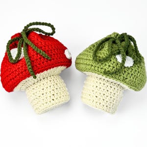 May include: Two crocheted mushroom-shaped pouches with drawstring closures. One pouch features a red cap with white spots, and the other has a green cap with white spots. Both have cream-colored stems and dark green yarn ties at the top.