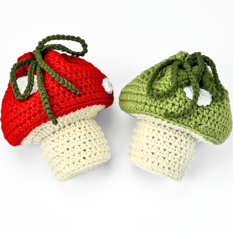 May include: Two crocheted mushroom-shaped pouches with drawstring closures. One pouch features a red cap with white spots, and the other has a green cap with white spots. Both have cream-colored stems and dark green yarn ties at the top.