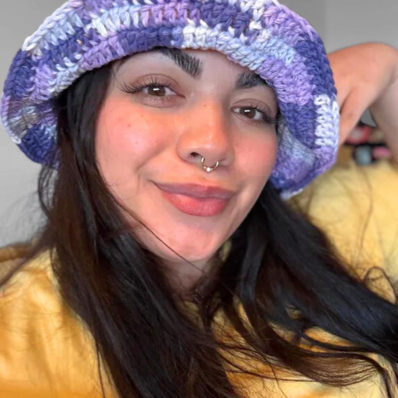 May include: A handmade crochet bucket hat featuring a variegated colour pattern of deep purple, light lavender, and white yarn. The hat has a wide, flexible brim and a textured, chunky knit construction with visible interlocking yarn loops throughout.