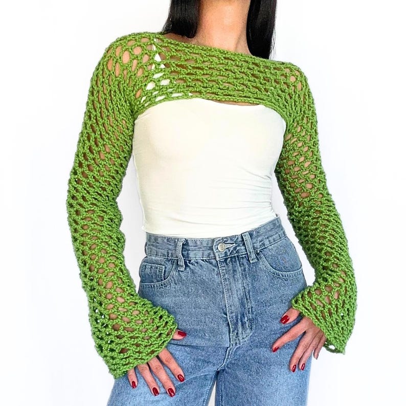 May include: A green crochet shrug with long sleeves and a wide, open neckline. The shrug is made with a delicate, open stitch pattern.
