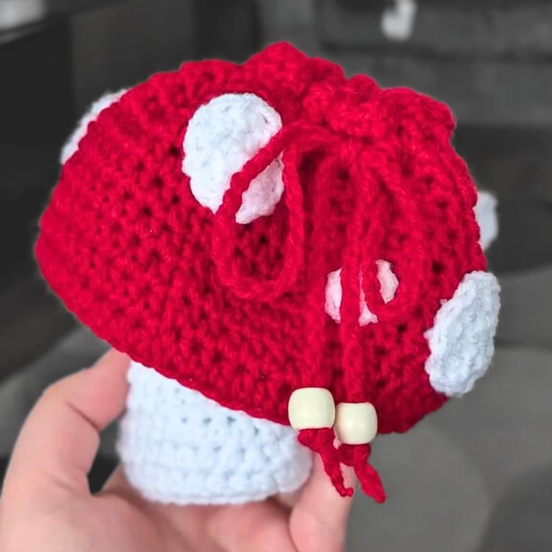 May include: A red and white crocheted mushroom-shaped drawstring pouch. The cap is bright red with white circular patches, while the stem is white. A red yarn drawstring closure features two light-colored wooden beads at the ends of the ties.