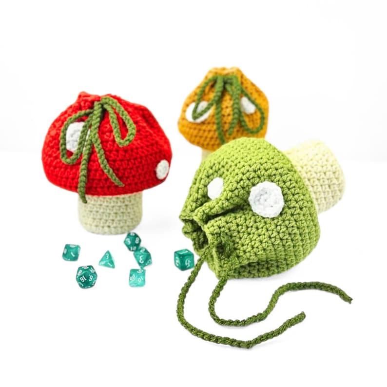 May include: Three crocheted mushroom-shaped drawstring pouches in red, yellow, and green, each featuring white spots and a light-colored stem. A collection of small, multi-sided green dice is scattered on the surface near the pouches.