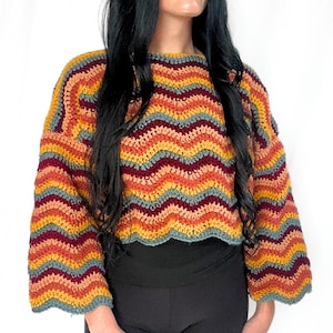 May include: A long-sleeved crochet sweater featuring a horizontal chevron pattern in alternating stripes of mustard yellow, burnt orange, deep maroon, and muted slate blue. The garment has a round neckline and a scalloped hemline at the waist.