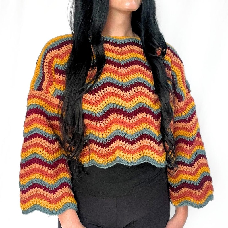 May include: A long-sleeved crochet sweater featuring a horizontal chevron pattern in alternating stripes of mustard yellow, burnt orange, deep maroon, and muted slate blue. The garment has a round neckline and a scalloped hemline at the waist.