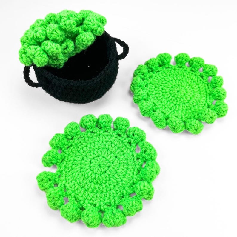 May include: A black crocheted cauldron with two side handles and a textured green crocheted lid, accompanied by two round, bright green crocheted coasters featuring a decorative bobble-stitch border around the circular edges.