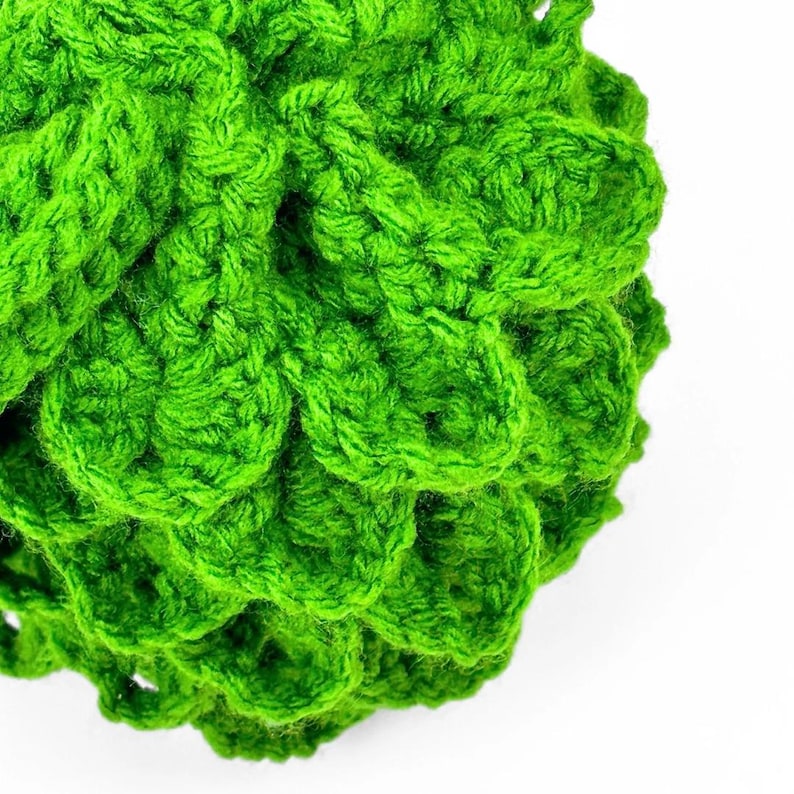 May include: A bright green crocheted sphere featuring a textured, layered scale-like pattern created with yarn. The object has a rounded, three-dimensional shape with visible interlocking stitches throughout the entire surface of the item.