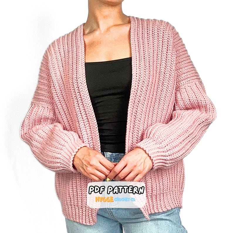 Op de afbeelding: Een lichtroze, gehaakt vest met lange mouwen en een open voorzijde, voorzien van een geribbelde textuur en verlaagde schouders. Het kledingstuk heeft een label onderaan in het midden met de tekst PDF PATTERN HYGGE CROCHET CO. in zwart en oranje.