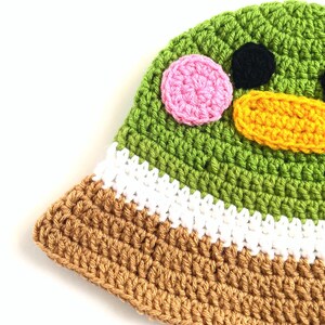 Pu&ograve; includere: Cappello a secchiello verde all'uncinetto con bordo marrone e bianco. Il cappello ha un viso da cartone animato con occhi rosa e neri e un becco giallo.