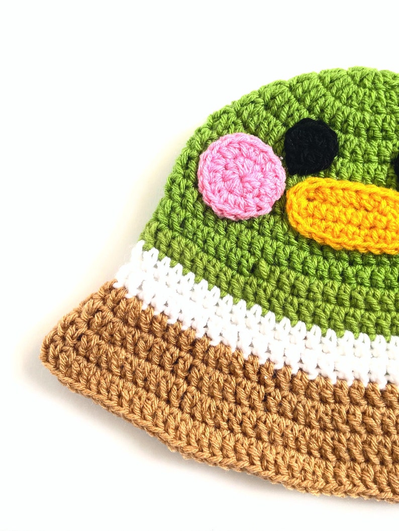 Pu&ograve; includere: Cappello a secchiello verde all'uncinetto con bordo marrone e bianco. Il cappello ha un viso da cartone animato con occhi rosa e neri e un becco giallo.
