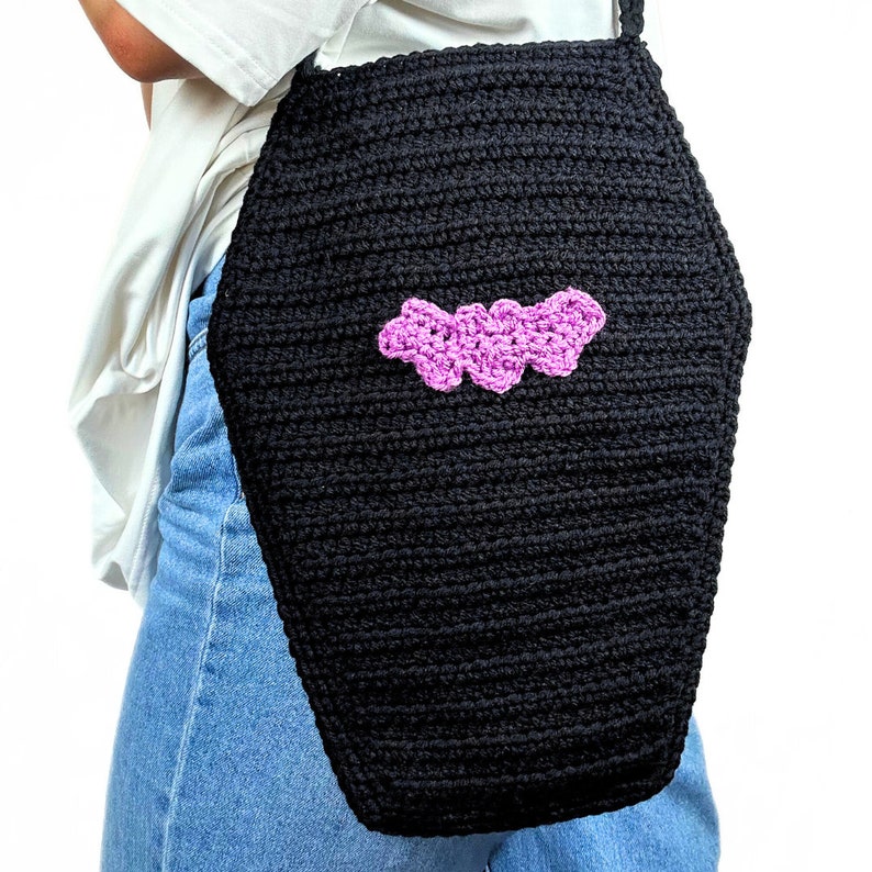 May include: A black coffin-shaped crochet bag featuring a pink crocheted bat appliqu&eacute; centered on the front. The bag is constructed with a dense, textured stitch pattern and includes a matching black crochet strap attached to the top corner.